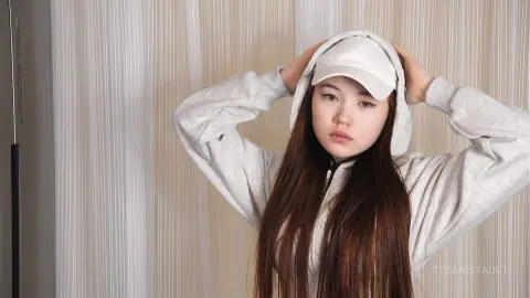 Young woman in a white hoodie and baseball cap posing against a neutral curtain background with a relaxed mood.