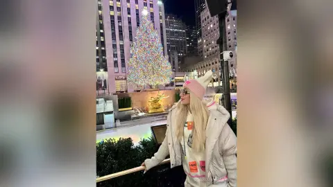 Blonde woman in a white puffer jacket and pink beanie smiling in front of a glowing Rockefeller Center Christmas tree.