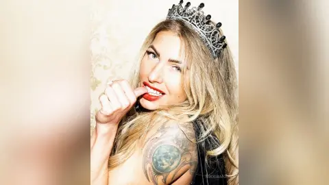 Smiling blonde woman with a silver crown and tattoos wearing a black outfit in a bright setting.