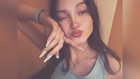 Young woman in a light blue tank top winking and blowing a kiss in a warm, cozy indoor setting.