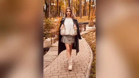 Young woman in a cozy white sweater and grey skirt walking along a scenic autumn park path.