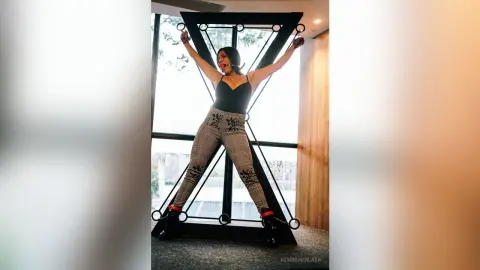 Confident woman in black bodysuit and patterned leggings posing with arms raised in a modern interior setting.