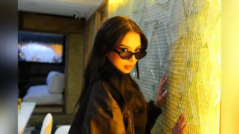 Stylish woman in a black leather jacket leaning against a glowing gold mosaic wall in a modern luxury interior.
