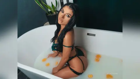 AlmaKane poses naked in a white bathtub filled with milky white water and orange slices, wearing dark green lace lingerie.