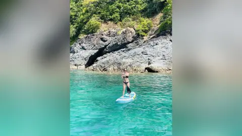 Woman in black bikini paddleboarding on turquoise ocean water near rocky cliffs on a sunny day.