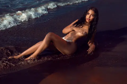 JasminMale poses naked on a dark sandy beach, wearing only a white bikini bottom while leaning back against the wet sand and waves.