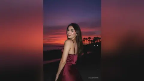 Elegant woman in a maroon satin slip dress standing against a dramatic sunset sky by the ocean.