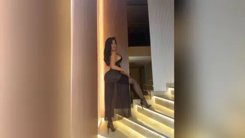 Elegant woman in a black sheer evening gown posing on a modern lit staircase in a luxury interior setting.