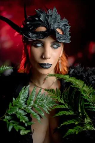 AdelinaVanz wears a black feathered masquerade mask and dark lipstick while posing with green ferns against a red background.