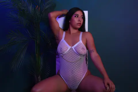 Young woman in white mesh bodysuit posing against a dark blue background with tropical palm leaves.