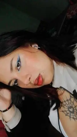 Close-up portrait of a woman with dark hair, blue eyeliner, and a spiderweb tattoo wearing a white top.