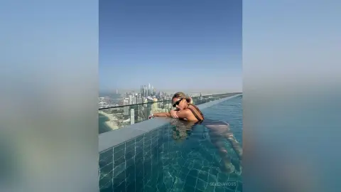 Stylish woman in a black bikini relaxing in a rooftop infinity pool overlooking a sunny city skyline.