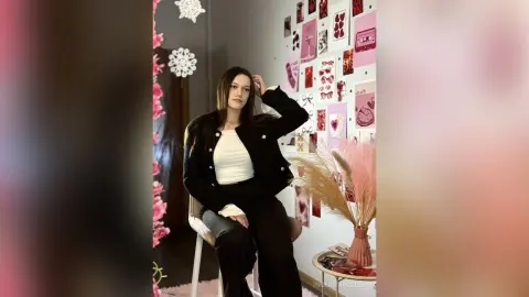 IngerMarzella sits in a chair wearing a black jacket and white top, posing with one hand in her long dark hair against a wall decorated with pink photos.