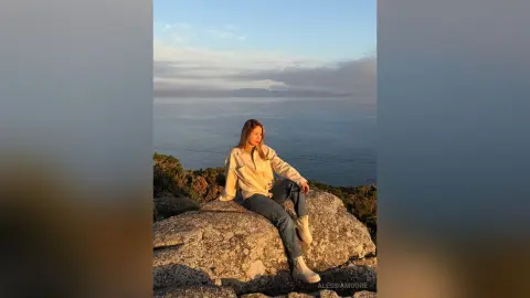 Young woman in a beige jacket and jeans sitting on rocks overlooking the ocean at sunset.