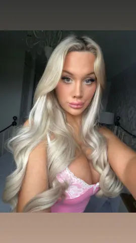 Blonde woman in a pink lace camisole posing with long wavy hair in a cozy bedroom setting.