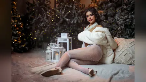 ZaraCastelli poses sitting up in white fishnet tights and a white furry coat, surrounded by lit Christmas trees and white lanterns.