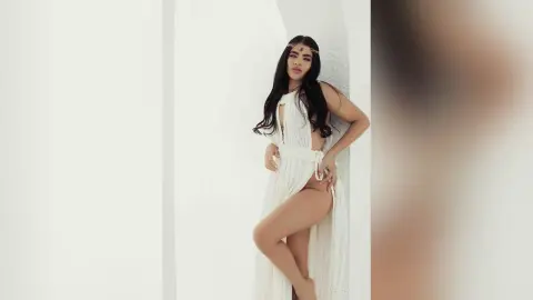 Elegant woman in a flowing white Grecian gown posing against a minimalist white wall in a dreamy aesthetic.