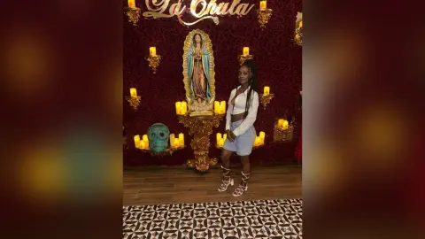 Young woman in a white crop top and denim shorts standing before a religious statue and glowing candles.