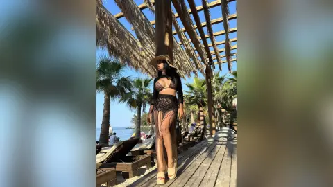 NicoleBella stands under a wooden pergola wearing a black lace bikini and a wide-brimmed sun hat near palm trees.