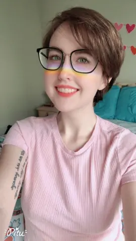 Smiling young woman with short brown hair and glasses wearing a pink ribbed t-shirt in a bright bedroom.