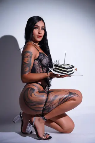 Woman in black lace lingerie with large leg tattoos holding a birthday cake in a studio setting.