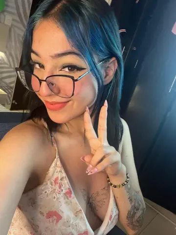 Smiling young woman with blue hair and glasses wearing a floral camisole making a peace sign indoors.
