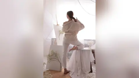 Woman in beige linen outfit sitting by a bright window in a minimalist, airy room with a serene mood.