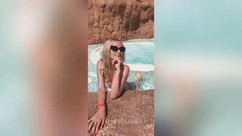 Blonde woman in a white bikini relaxing in a turquoise pool against a desert rock background on a sunny day.