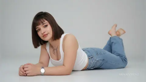 Young woman with short brown hair in a white tank top and blue jeans lying on a white floor, relaxed mood.