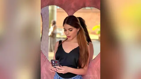 ValeryRouge wears a black v-neck camisole and blue jeans while looking down at her phone outdoors.