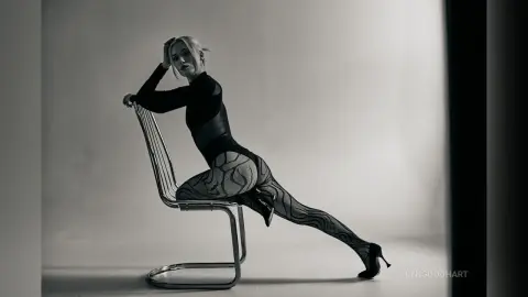 Elegant woman in a black bodysuit and patterned tights posing on a silver chair in a minimalist studio setting.