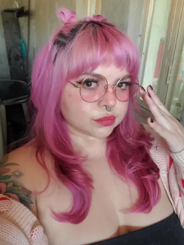 Woman with pink hair and round glasses wearing a cozy pink cardigan in a bright indoor setting.