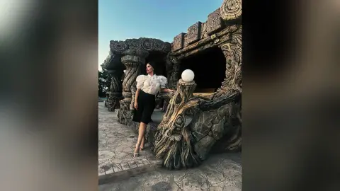 AnnaKarev poses in a white puff-sleeve blouse and black shorts against a large, carved stone structure.