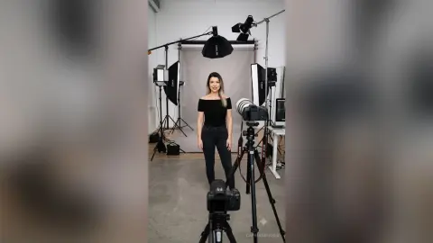 CapriAndersen stands in a studio setting wearing a black off-the-shoulder top and black jeans, surrounded by professional lighting and camera equipment.