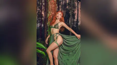 LaurentBelluci poses in a green bikini and long flowing skirt against a forest background, showing her long red hair and bare legs.