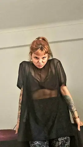 Young woman with red hair wearing a sheer black mesh top and dark skirt in a minimalist indoor setting.