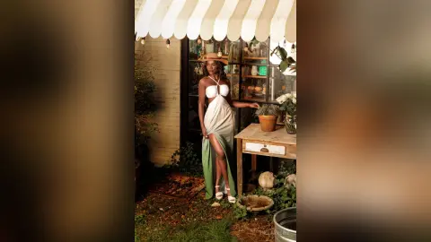 Elegant woman in a white and green flowing dress posing by a rustic outdoor cafe table in soft sunlight.