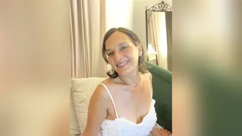 MichaelaBelle smiles while wearing a white ruffled camisole, sitting against a green velvet chair.