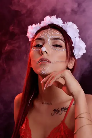 Woman in red lace outfit with white spiderweb face paint and floral crown against a moody dark background.