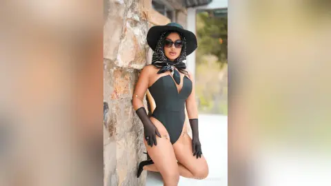 Stylish woman in a black bodysuit and wide-brimmed hat posing against a stone wall in bright sunlight.