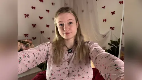 Young woman in a pink floral cardigan smiling in a cozy room with white curtains and red decor.
