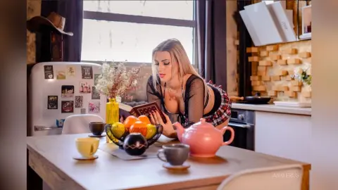 Blonde woman in a black mesh top reading a book at a dining table with fruit and tea in a bright kitchen.