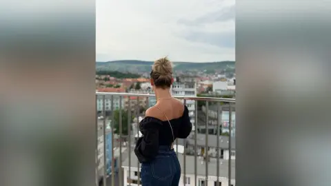 SelenaRusse stands on a balcony overlooking a city, wearing a black off-the-shoulder top and blue jeans.