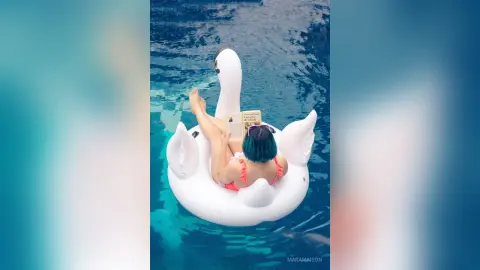Relaxed woman in a pink bikini reading a book on a white swan floatie in a blue swimming pool.