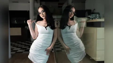 MayaOllin poses in a tight white dress, looking at the camera with long dark hair and glasses in a room with a checkered floor.