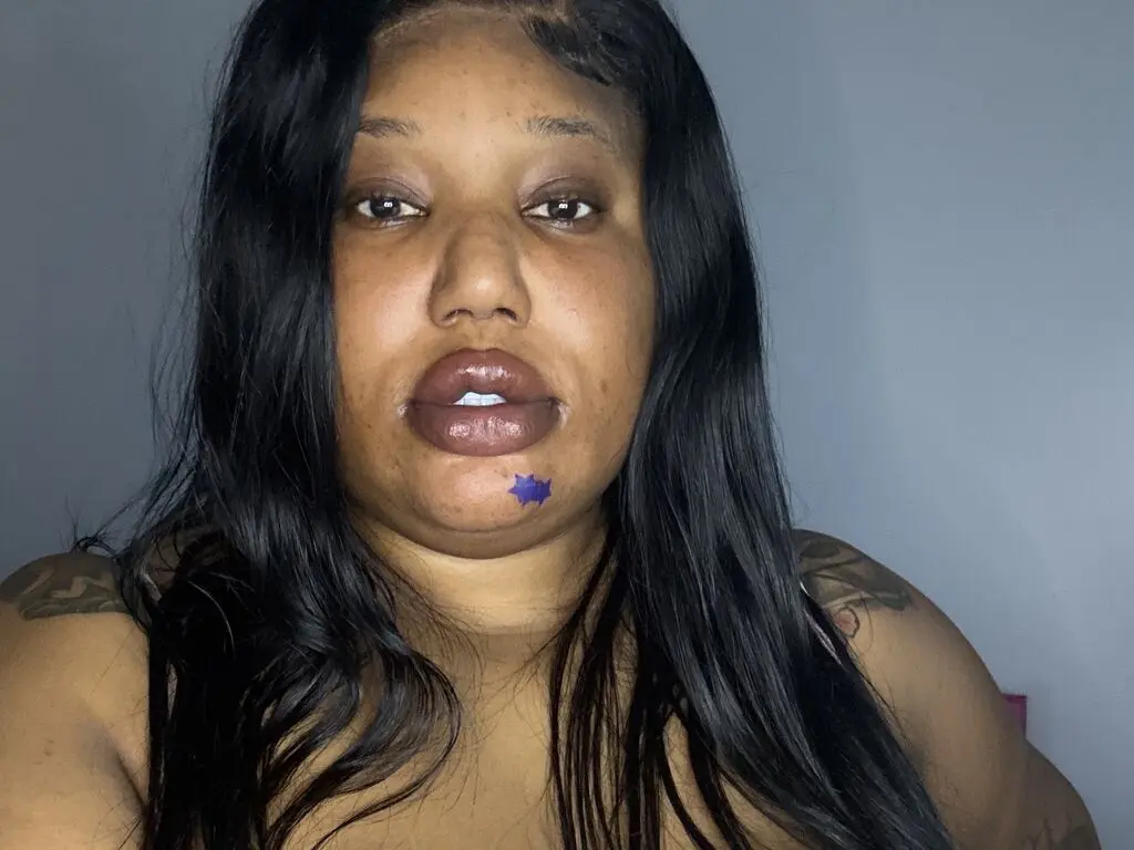 FATBIGBBW looks into the camera with long black hair and a purple star tattoo on her chin, showing her bare shoulders and chest.