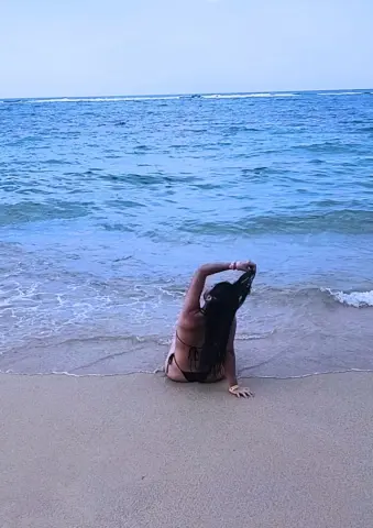 Young woman in a black bikini sitting on a sandy beach facing the blue ocean waves under a clear sky.