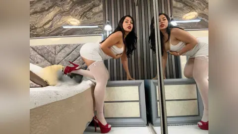 Woman in white mini dress and white thigh-high stockings posing on a bed in a modern luxury bedroom.