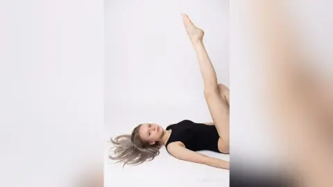 SophiePeachy lies on her back on a white floor, wearing a black bodysuit with her legs raised high in the air.