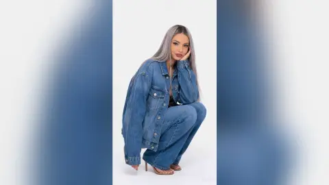 Kalira poses in a denim jacket and matching jeans, leaning her head on her hand against a white background.